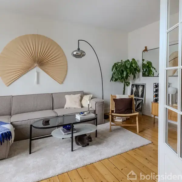 En stue med en grå sofa med puder og et blåt tæppe. En buet lampe står ved siden af, og en stor, grøn plante er i hjørnet.