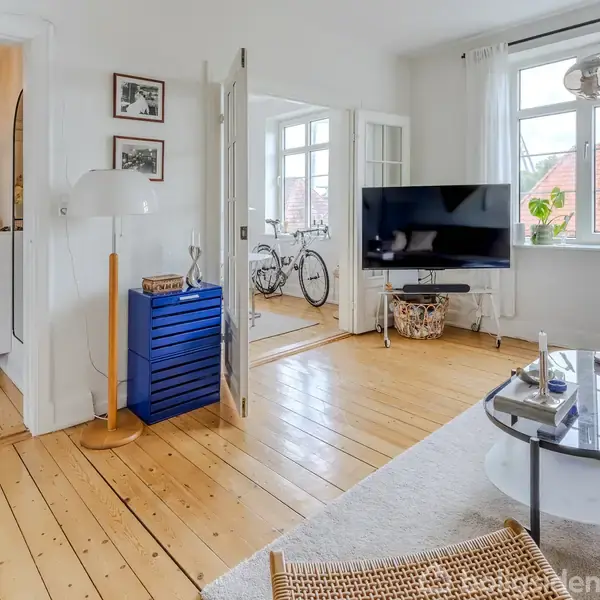 Stue med træplankegulv og moderne møbler; tv er monteret foran et stort vindue. En cykel ses gennem franske døre i næste rum. Entreen til venstre har en hængende lampe.