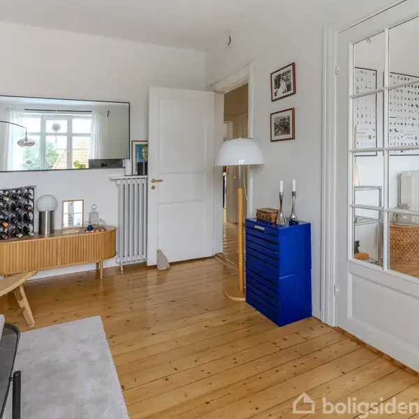 Stue med trægulv og moderne møbler. Sofa ved siden af et glasbord. Blå skuffemøbel ved døren, spejl og planter på væggen. Spisebord i baggrunden. Ingen synlig tekst.
