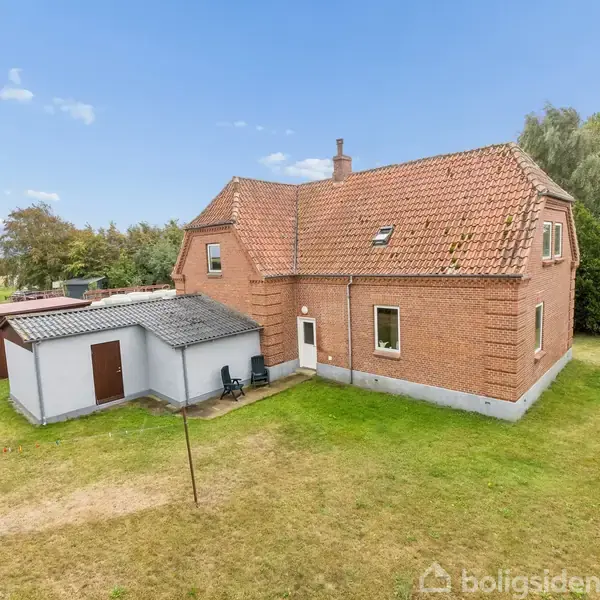 Et rødt murstenshus med rødt tegltag står i en grøn have. Ved siden af huset ligger en hvid tilbygning med to stole foran. Omgivelserne er landlige med træer.
