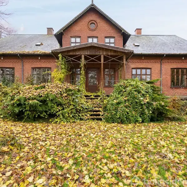 En murstensvilla står stille, delvist dækket af efterårsblade og vildtvoksende buske. Huset har et spidstag og flere vinduer. I forgrunden er en græsplæne dækket af nedfaldne blade.