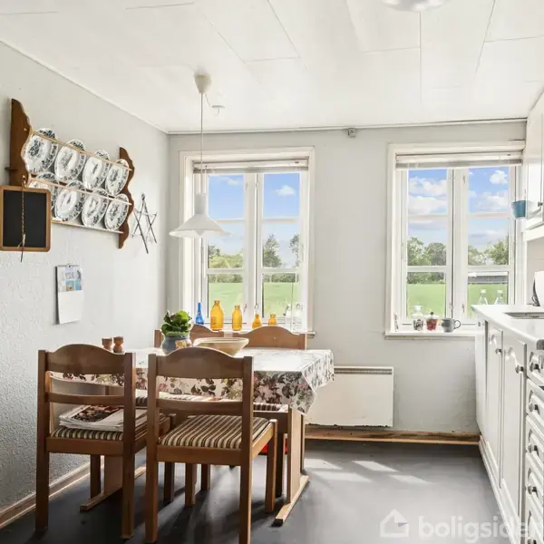 Et køkken med træmøbler står ved et bord med dug. Der er tallerkener hængt på væggen. Store vinduer giver udsigt til en grøn mark udenfor.