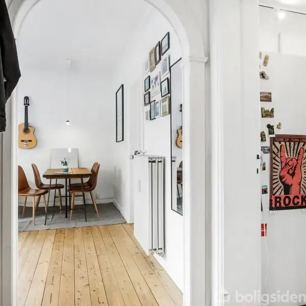 Guitar hænger på hvid væg ved spisebord og stole, omgivet af billederammer og radiator. Til højre ses køleskab med en "ROCK" plakat i et køkkenområde med kasser og fliser.