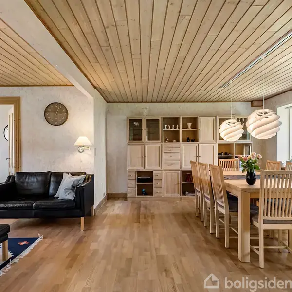 En hyggelig stue med et sort lædermøblement, en spisebord med seks træstole. Der er trægulv og træloft, to moderne lamper hænger over bordet. Reol med opbevaring i baggrunden.