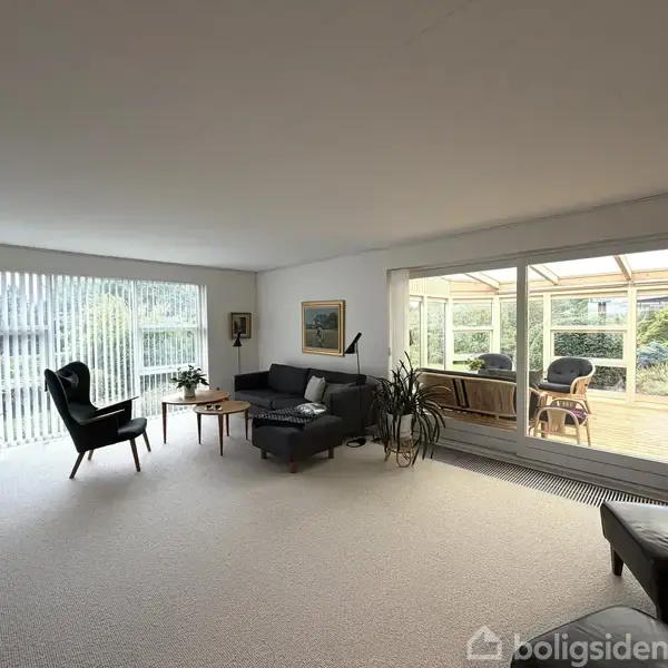 En stue med en mørkegrå sofagruppe og lænestol, står tæt ved et stort vindue med persienner, giver udsigt til en møbleret terrasse i sollys. Ingen tekst fundet.