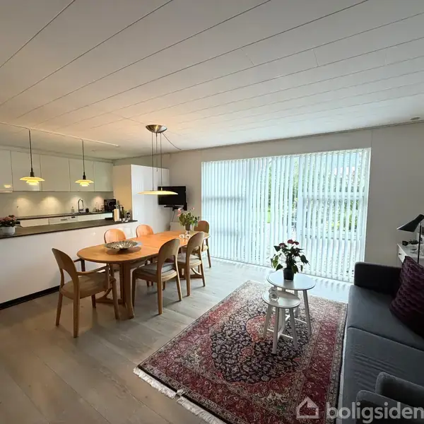 Spisestue med træbord og stole, dekoreret med en frugtskål, beliggende ved siden af åbent køkken med ophængte lamper. Stuen indeholder sofa, tæppe og borde ved glasdør med lameller.