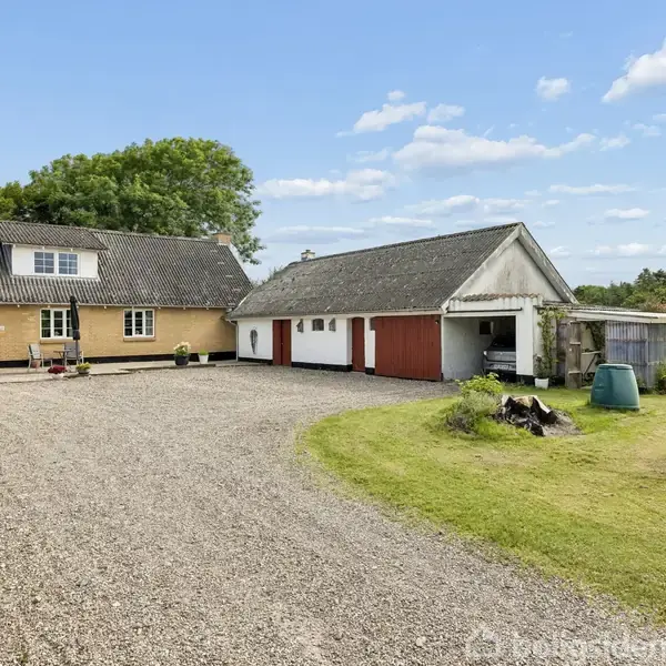 Gulstenshus med skråtag står foran en grusbelagt gårdsplads. Ved siden af ses en hvid garage med røde porte. Græsplæne og træer omgiver området under en blå himmel.