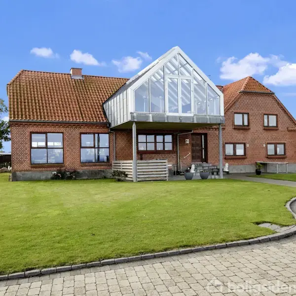 Et rødt murstenshus står stille i en velplejet have med grøn græsplæne og blå himmel. En stor glasveranda hænger over indgangen, omgivet af flisebelagt indkørsel og få træer.