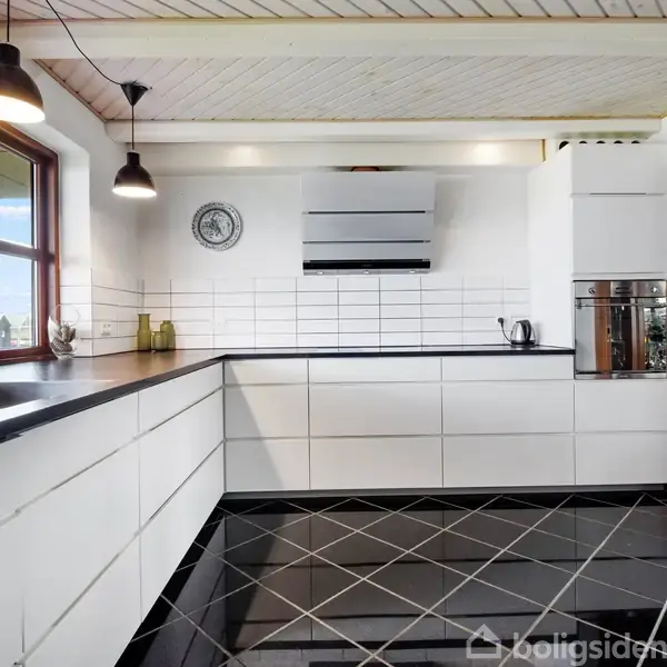 Moderne køkken med hvide skabe og sorte bordplader i et L-formet layout. Store vinduer giver naturligt lys. Sort-hvidt flisegulv og indbygget ovn. Loftslamper hænger fra loftet.
