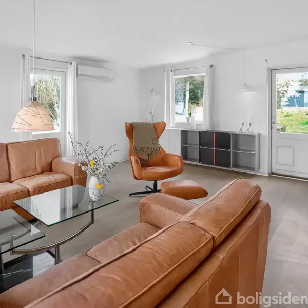 Brun lædersofa og lænestol står i en lys stue med glasbord. Store vinduer og dør åbner ud til en grøn have. Baggrunden har et skab og minimalistisk indretning.