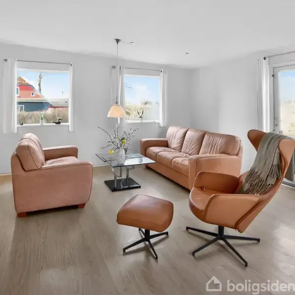Stue med lænestole og sofa i læder står omkring et sofabord på et trægulv. Et vindue og en glasdør giver udsigt til en have.