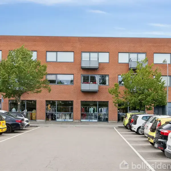 En murstensbygning med store vinduer står stille. Parkeringspladsen foran er fyldt med biler. Omkring bygningen er der træer, og der er en blå himmel i baggrunden.