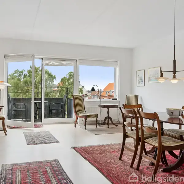 Et spisebord med stole står i en lys stue med træmøbler og tæpper. Glasskydedøre åbner til en balkon med udsigt til grønne træer og et rødt hustag.