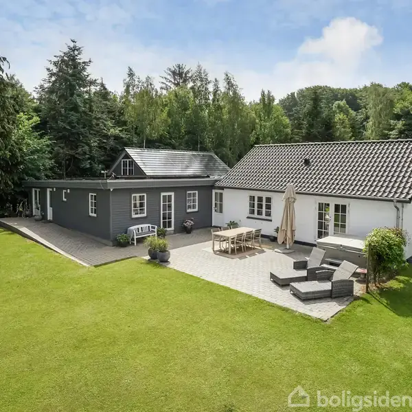 Et enkelt plan hus med hvide og grå facader står ved skovgrænse. Terrassen har et spisebord og sofaer under en parasol, omringet af en grøn græsplæne.