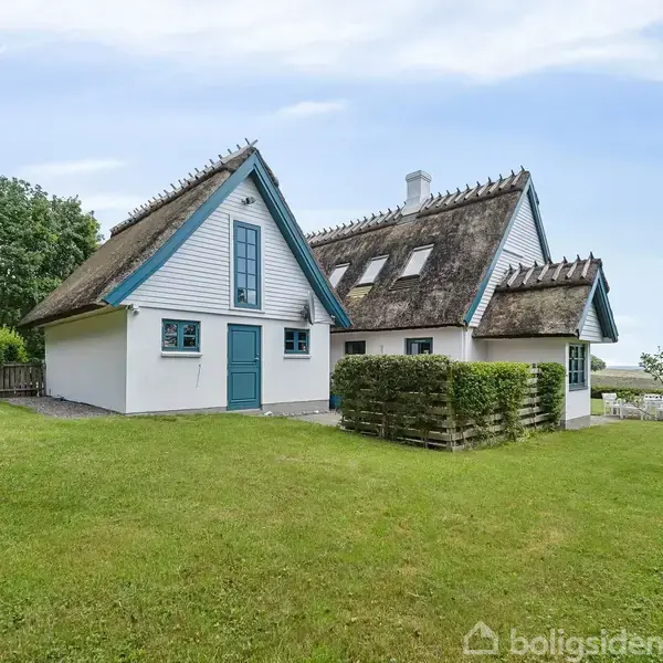 Stråtækt hus med blå døre og rammer står på en grøn græsplæne. I baggrunden er der træer og udsigt til havet. En rød vandhane står til venstre.