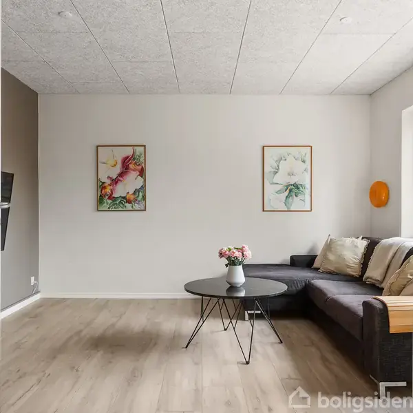 Et sort sofabord med blomster på står foran en mørk sofa i en lys stue med trægulve og kunstværker på væggen.