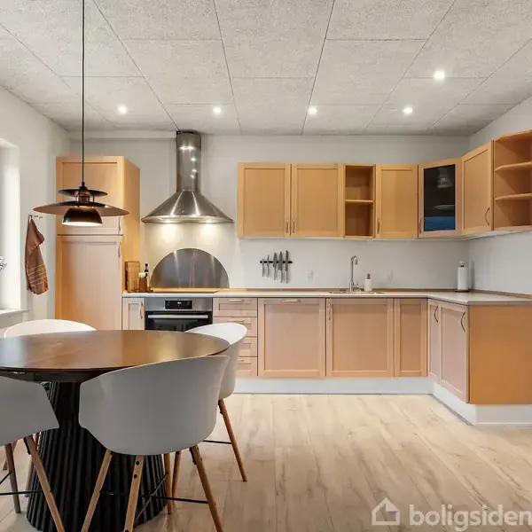 Et lyst køkken med træskabe og stålventilator. Et rundt spisebord med fire hvide stole står i midten. Store vinduer lader naturligt lys strømme ind.