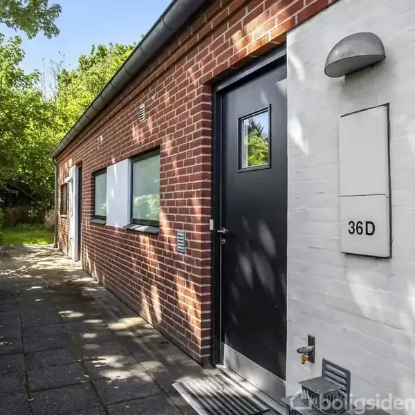 En murstensbygning med en sort dør markeret med "36D". En flisebelagt sti løber langs siden. Omgivet af grønne træer og buske.