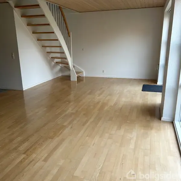 Trætrappe fører opad i hjørnet af et tomt rum med trægulv og hvide vægge. Store vinduer slipper naturligt lys ind fra højre side.