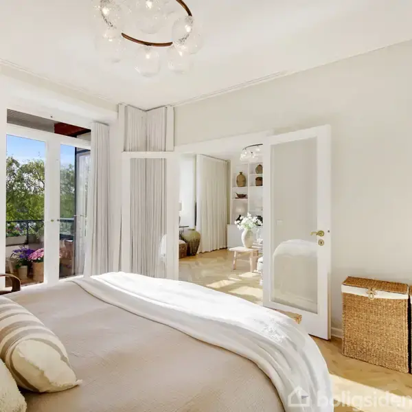 Et soveværelse med en dobbeltseng dækket af hvide og beige sengetæpper, åbner gennem franske døre til en balkon med udsigt til træer. To kurve står ved væggen.
