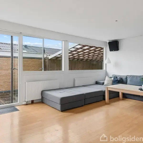 En grå hjørnesofa står i et lyst rum ved en glasdør. Der ligger en træbord foran sofaen, og udenfor ses en terrasse med overdækning. Væggen er hvid med en højttaler monteret.