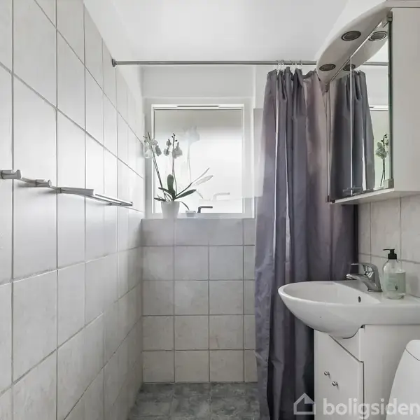 Håndvask med spejl hænger på væggen, omkranset af hvide fliser. En grå bruseforhæng ses ved siden af, og der er et vindue med en orkidéplante. Ingen tekst til stede.