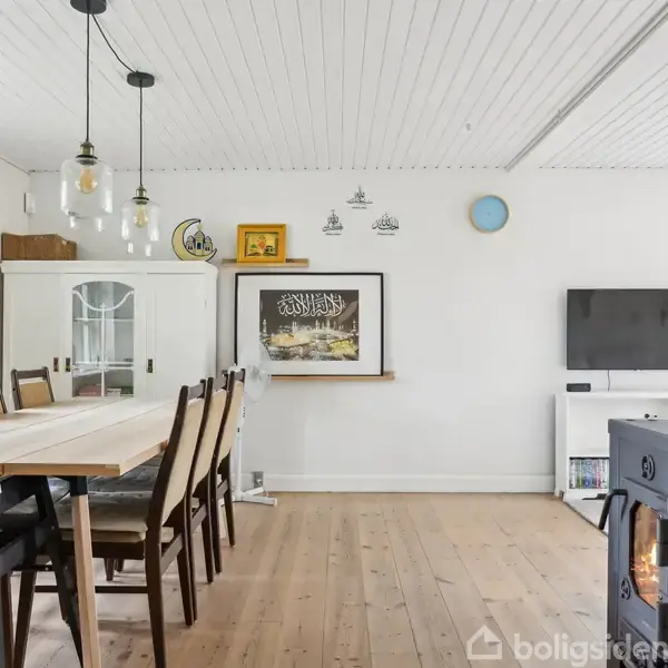 Et spisebord med flere stole står i et lyst rum med hvide vægge og træloft. Der er kunst på væggen, en reol til højre, og en brændeovn ved siden af.