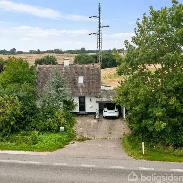 Hus med carport og bil parkeret i indkørslen, omkranset af træer og buske. Vejen foran huset, åbne marker og en elledning i baggrunden under en klar himmel.
