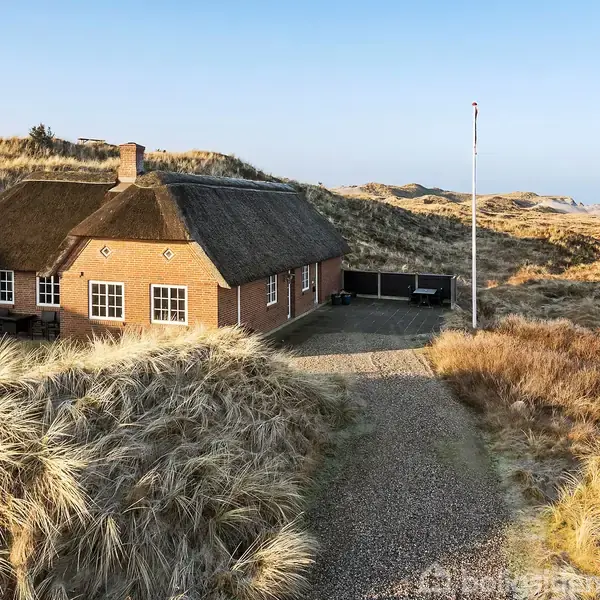 Et stråtækt hus står stille omgivet af sandede klitter og spredt græs. En grusvej fører op til husets indgang, og en flagstang står ved siden af.