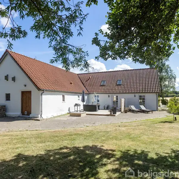 Hvidt landhus med rødt tegltag og trædør står på en solrig dag. To liggestole og grill ses på terrassen. Huset er omgivet af græs og træer under en blå himmel.