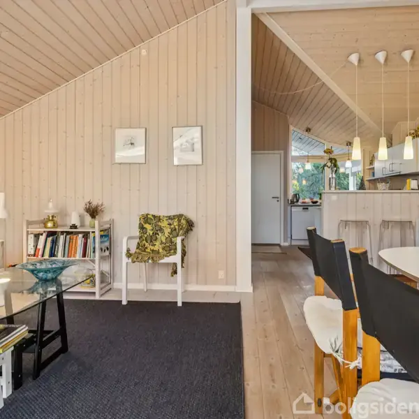 Stue viser glasbord med planter, omgivet af lys sofa og stole. Trævægge og loft giver hygge. Baggrunden viser køkken med bar og hængende lamper. Bogreol til venstre.