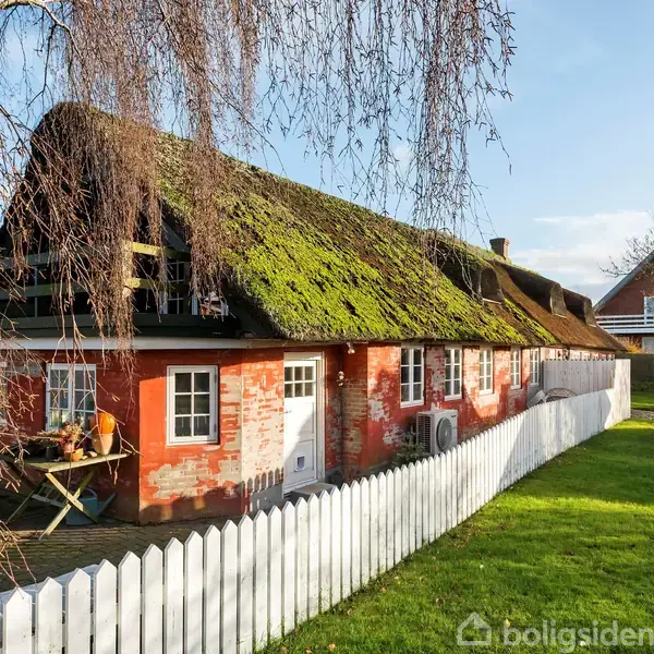 Et stråtækt hus med røde murstensvægge står omgivet af en hvid, træhegn i en grøn have med træer. Sollys skinner på taget, og en dansk flagstang ses i baggrunden.