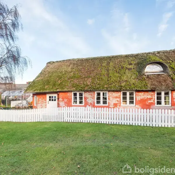 Gammelt bindingsværkshus med stråtag og runde vinduer står stille bag en hvid stakit. Grøn græsplæne i forgrunden og et bladløst træ til venstre giver naturlig omgivelse.