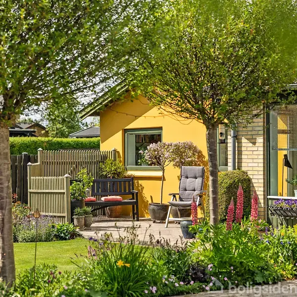 Et gult hus omgivet af en velholdt have med blomster og træer. En bænk og stol står på en terrasse foran huset, omgivet af en grøn, frodig græsplæne.