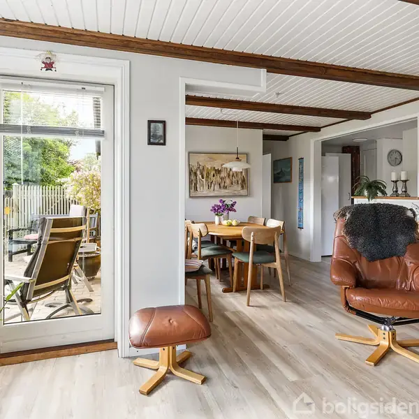 Lænestol i brun læder står i en stue med trægulv. Flere planter, en lampe og en spisebord med stole er synlige, og en dør fører ud til en terrasse.