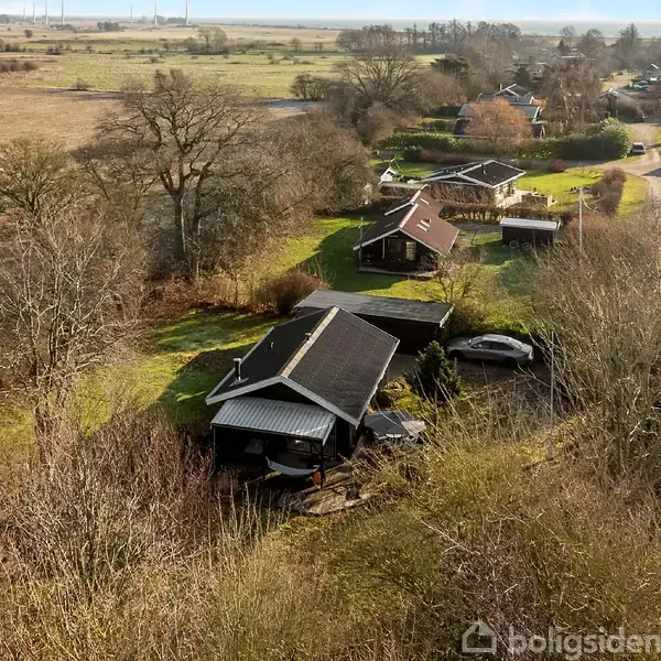 Hus set fra luften omgivet af træer og marker i et landligt område med flere huse i nærheden. En vej slingrer gennem landskabet.