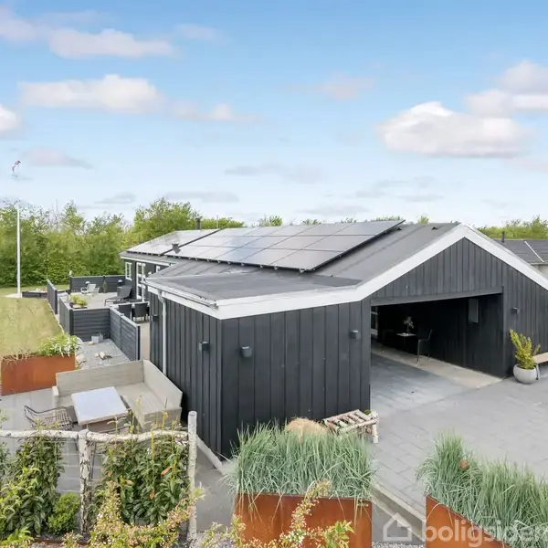 Et moderne sort træhus med solpaneler på taget, står i en velplejet have med terrasse, blomsterkasser og græsplæne. Husets garage er åben med en vej der fører ind.