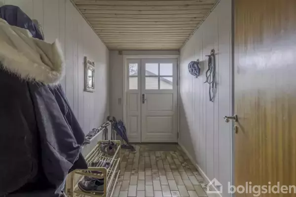 En entre med en dør i enden. Jakker hænger til venstre, og sko står på en skohylde. Vægge og loft er træbeklædt. En spejl hænger på væggen.