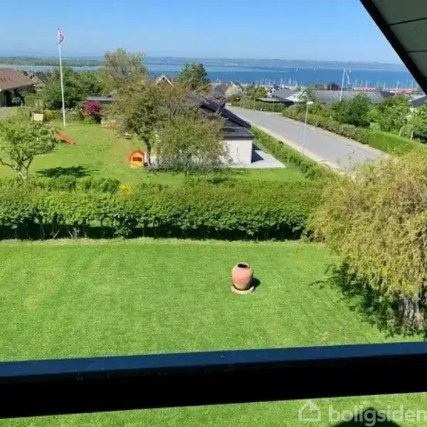 En frodig have med en lerkrukke i midten, grænset af hække. I baggrunden ses veje, huse og en fjord med både. Det er en solrig dag med klar himmel.