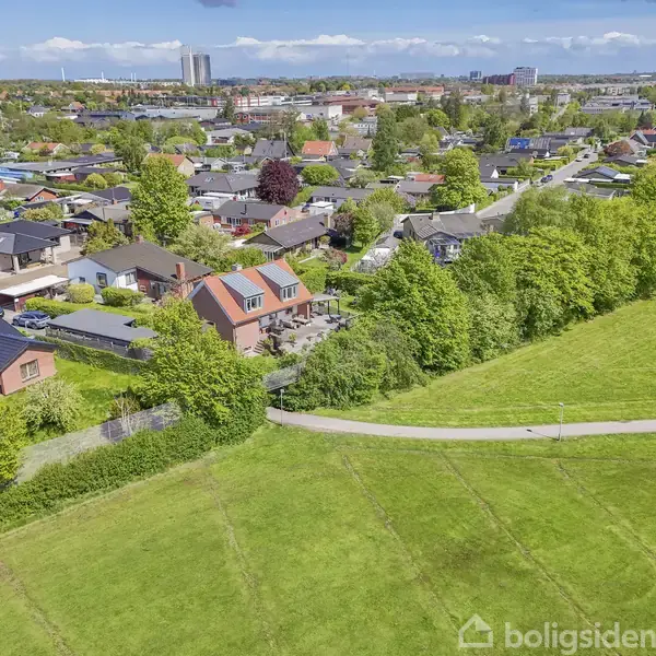 Huse står langs en park med grønne plæner og gangstier. Baggrund viser en by med spredte højhuse og trætoppene rækker over villaområderne.