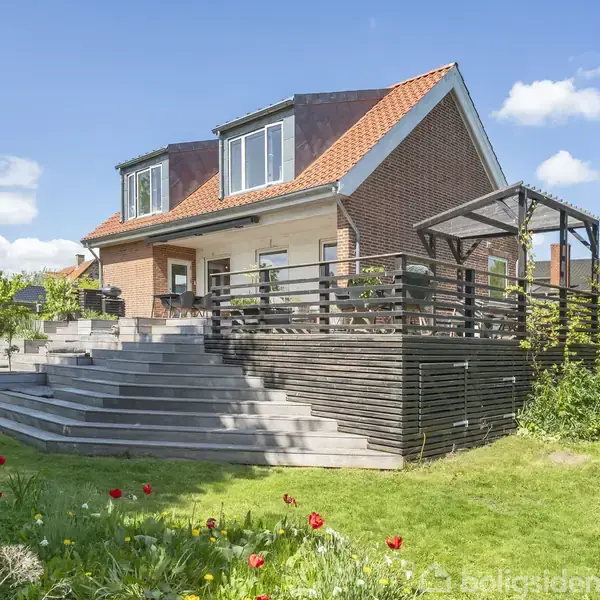 Hus med rødt tag står i en have. Store trappetrin fører til en hævet terrasse med gelænder. Græsplæne og blomster i forgrunden. Klar blå himmel i baggrunden.