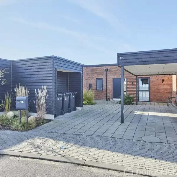 Hus med murstensvægge og carport i forgrunden, nummereret "15". Omkranset af flisebelagt område med affaldsbeholdere og få planter omkring. Skyfri himmel i baggrunden.