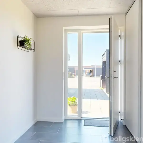 En åben dør med glasrude står på klem i en lys gang. Indendørs er hvide vægge og gråt flisegulv. Udenfor ses klar himmel og en gårdhave. En plante hænger på væggen.