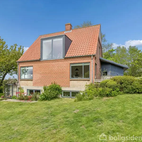 Hus med beige facade og rødt tag står ved siden af en garage med bil, i landlig omgivelser med marker og træer. Fortov med tre skraldespande foran huset.