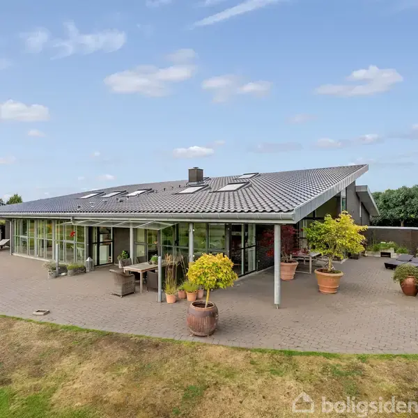 En moderne bungalow med store vinduespartier og gråtag belagt med solpaneler, omgivet af terrasse med krukker og grønt område. Skyer pryder himlen på en klar dag.