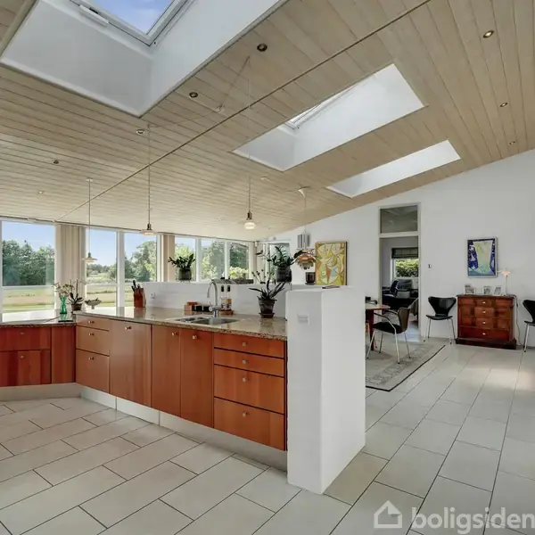 Køkkenet har et åbent layout med træloft og ovenlysvinduer. Køkkenøen er centrum og rummer blomsterpotter. Store vinduer giver udsigt over en grøn have.