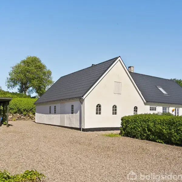 Hvidt hus med sort tag står i sollys, omgivet af grusvej og grønne træer. Til venstre holder en bil under en carport.