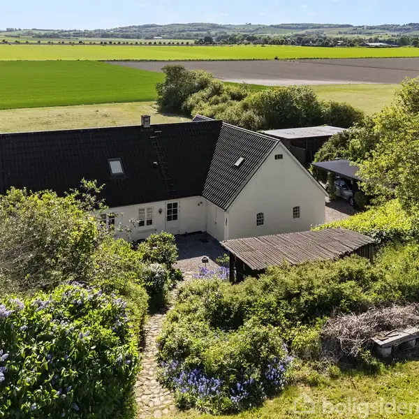 Gårdhus med sort tag står stille midt i frodig landlandskab. Omgivet af blomstrende buske, grønne marker og en lille terrasse, strækker naturen sig ud til horisonten i sollys.