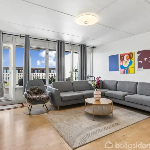 Gråt sofaarrangement står i et lyst stueområde med glasdør til balkon. Kunst på væggen og grønne planter skaber en hyggelig atmosfære. En stol og et rundt sofabord supplerer indretningen.