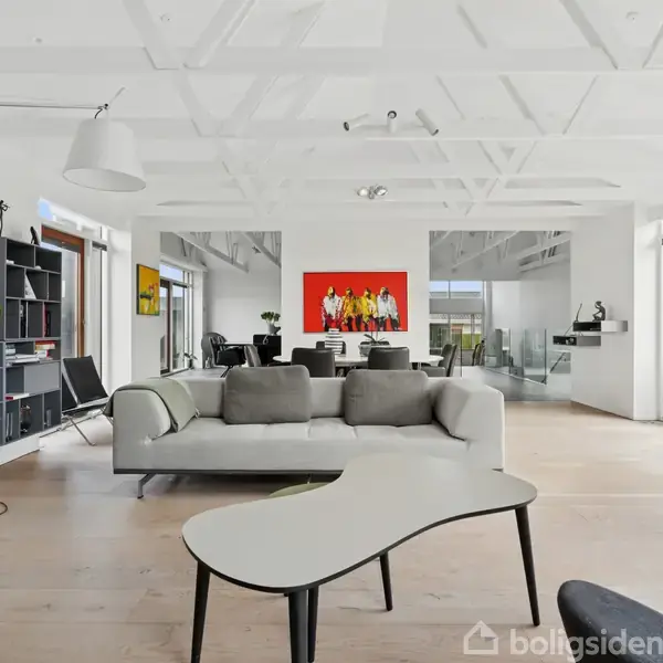 Stue med moderne sofa placeret foran et sofabord, omgivet af store vinduer med udsigt til haven. Hylder med dekorative genstande langs væggen. Maleri hænger over spisebordet.